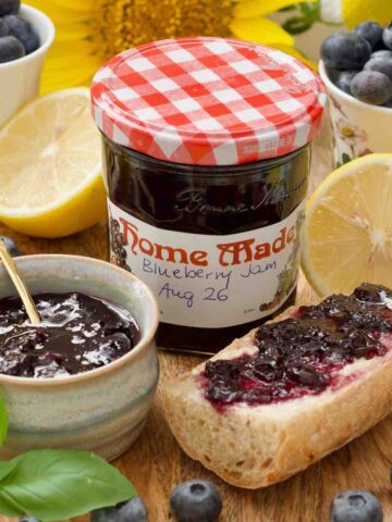 Close up of labelled blueberry jam jar, jam in a bowl and a piece of jam topped baguette.