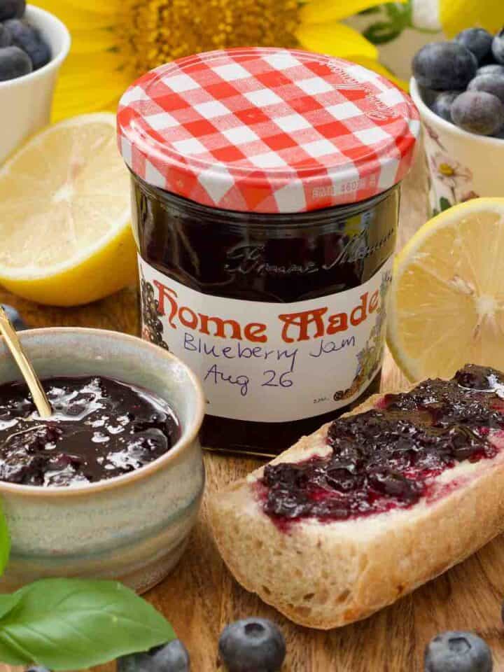 Close up of labelled blueberry jam jar, jam in a bowl and a piece of jam topped baguette.