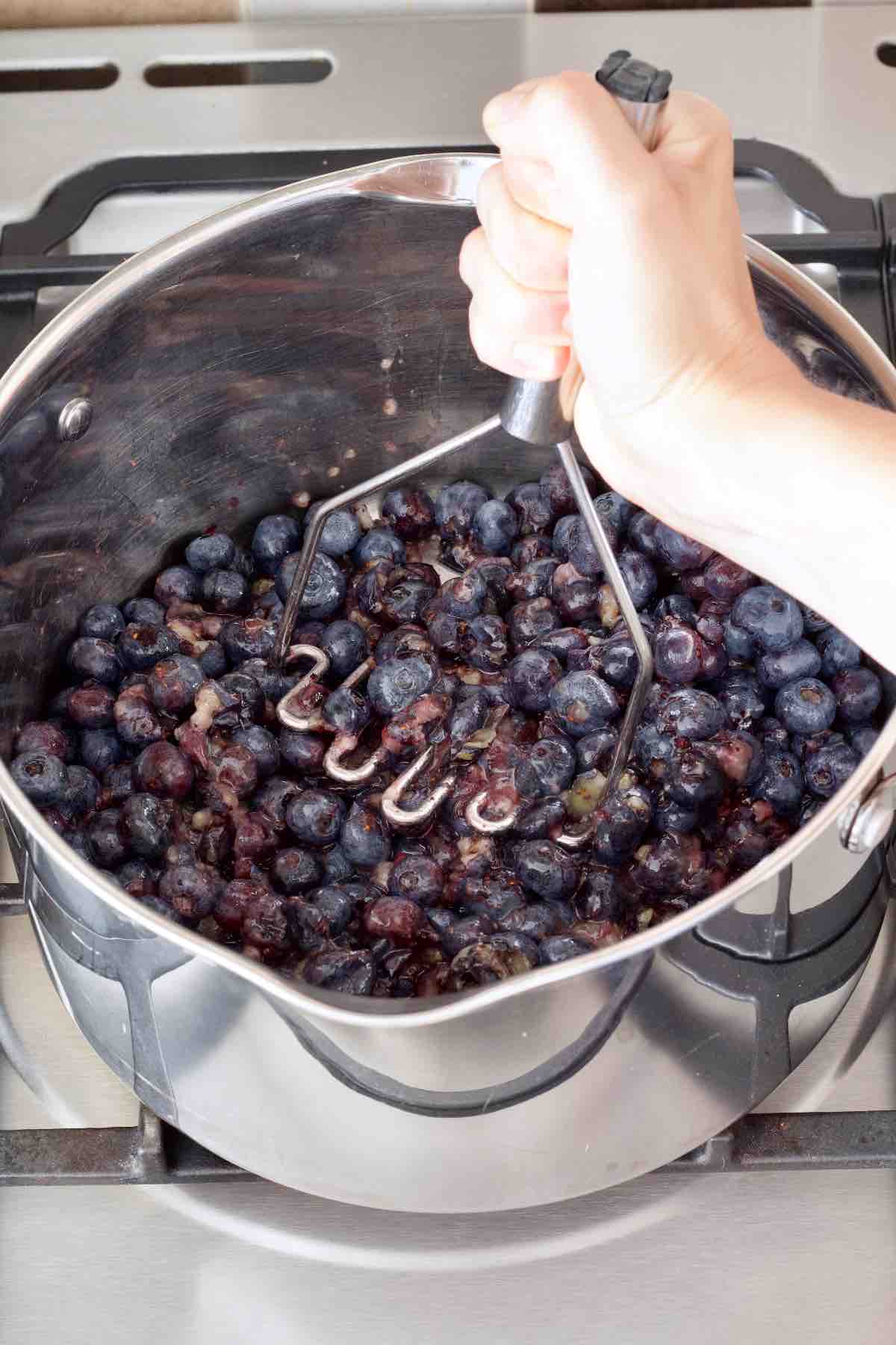 Mashing blueberries with potato masher in a pan.