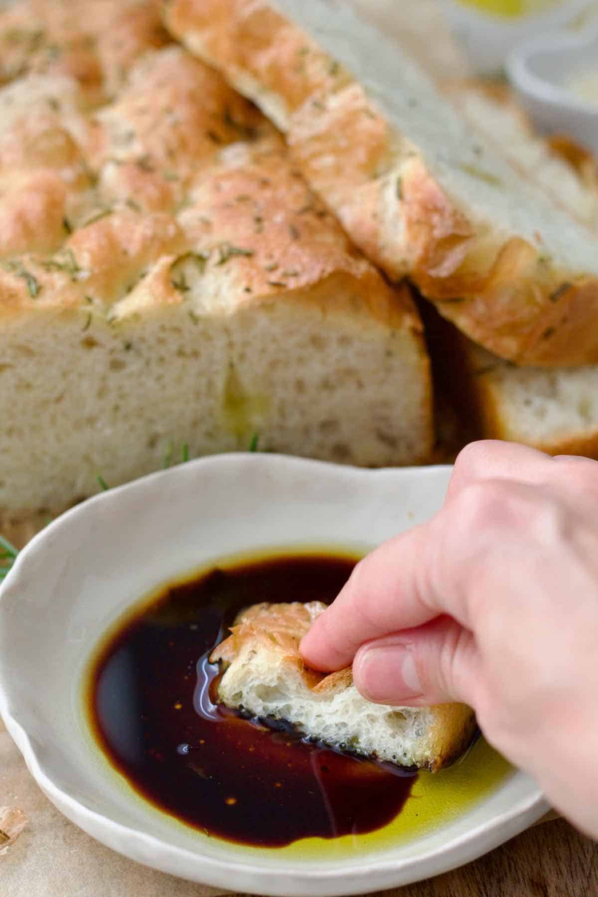 Hand dipping piece of focaccia in olive oil and balsamic vinegar dip.