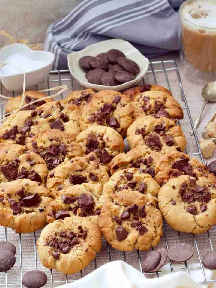Baked chocolate chip cookies on cooling rack with coffee, and chocolate buttons.