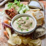 Red lentil hummus served in a bowl on a board with crisps, carrots, bread and more.