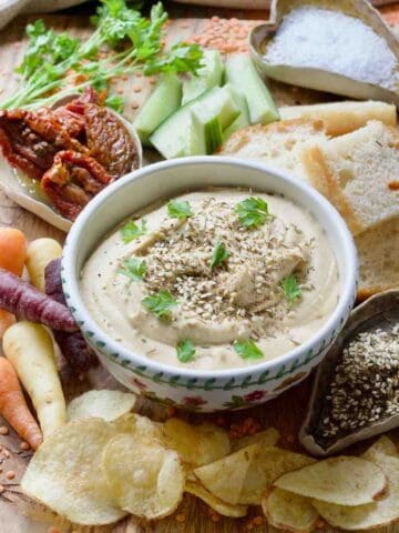 Red lentil hummus served in a bowl on a board with crisps, carrots, bread and more.