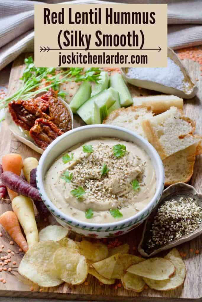 Bowl with red lentil hummus surrounded with snacks and seasonings.